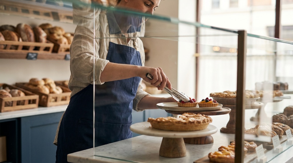 A baker using tongs to arrange pastries and pies in a glass bakery display cabinet